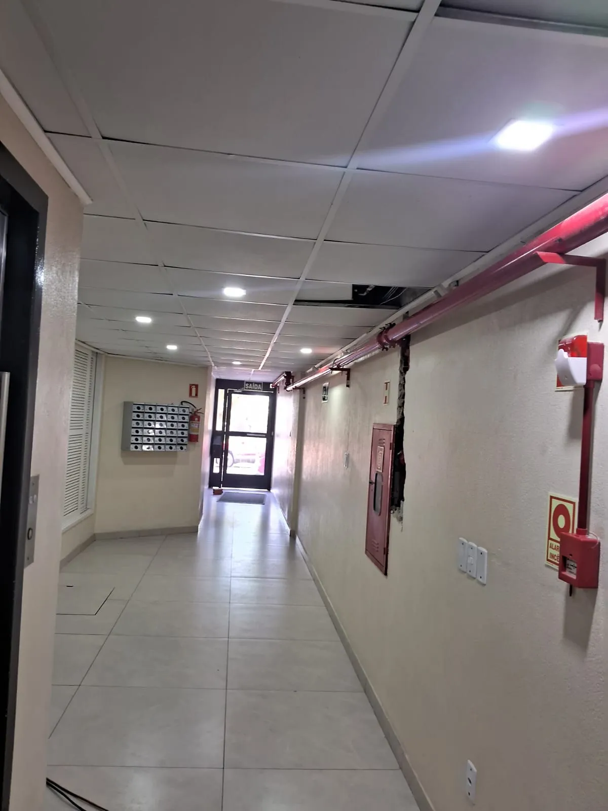Corredor de prédio com tubulação de combate a incêndio aparente, abrigos de mangueira e sinalização de saída